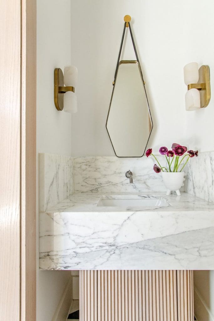Bathroom vanity with Carrara marble sink, geometric mirror, wall sconces, and a vase of flowers.