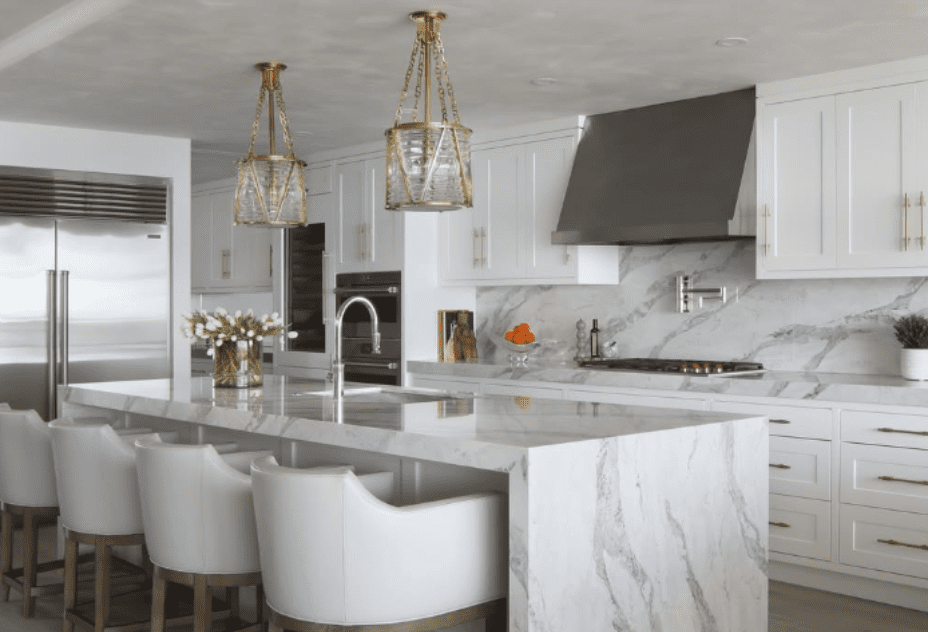 Luxurious kitchen with Pompeii Quartz Serenata/ Vicostone Quartz Amarcord waterfall island, white cabinetry, and gold pendant lighting.