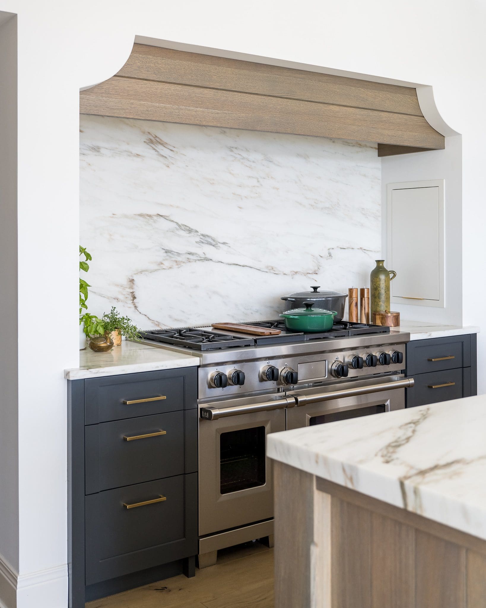 Modern kitchen with danby marble countertop and full-height backsplash behind stainless steel range, dark cabinetry, and wood range hood.