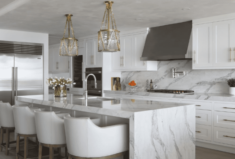 Luxurious kitchen with Pompeii Quartz Serenata/ Vicostone Quartz Amarcord waterfall island, white cabinetry, and gold pendant lighting.