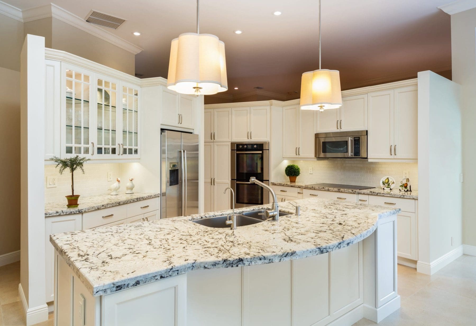 Traditional white kitchen with granite countertops, curved island, and drum pendant lights over a brown ceiling. 